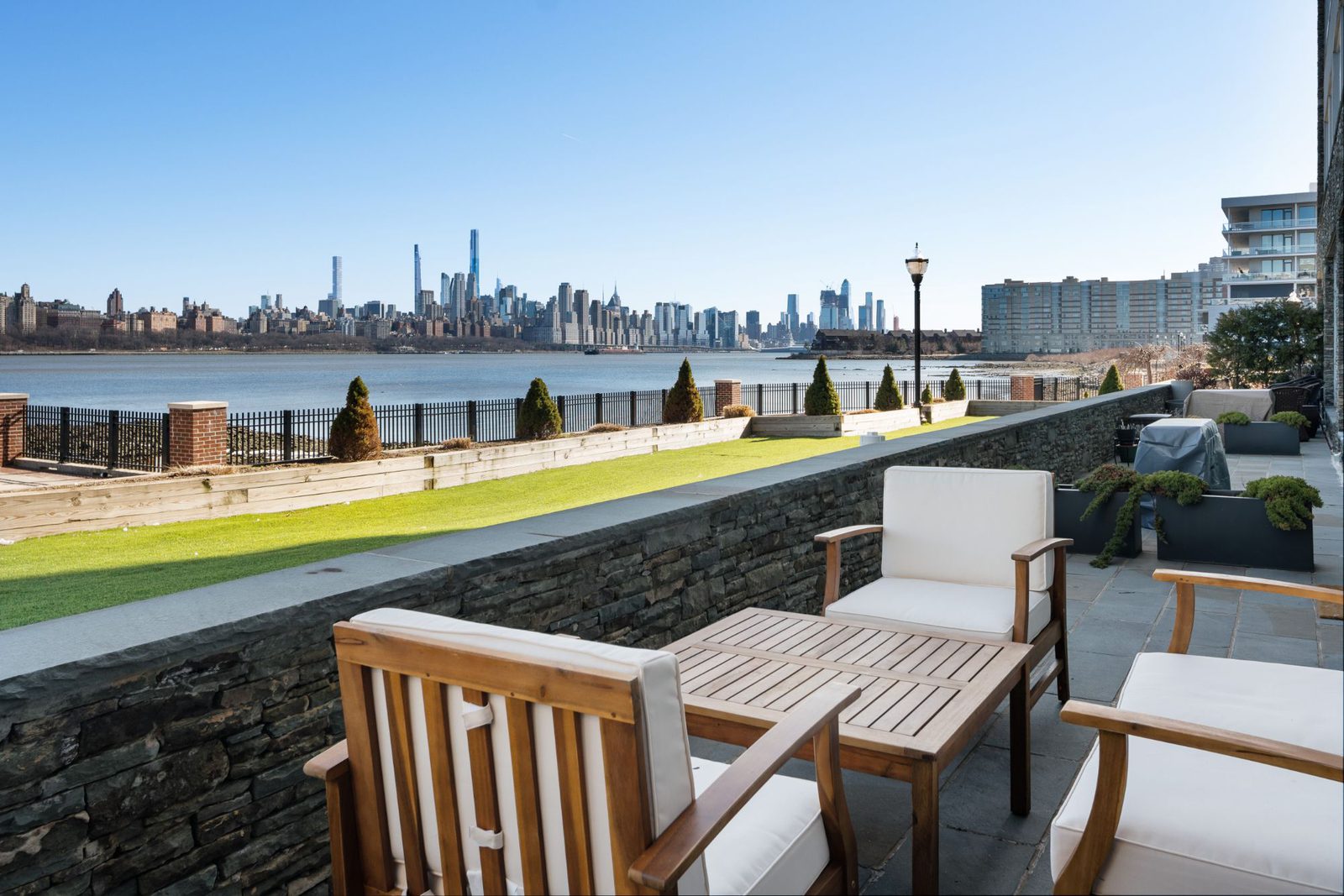 Private patio with wooden chairs beside a lawn on the Hudson River, with the Manhattan skyline across the water